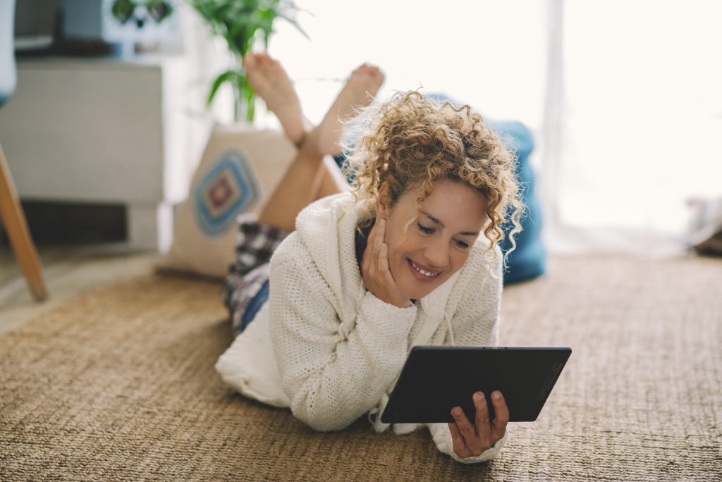 Happy adult woman laying on the carpet floor at home using modern tablet and internet connection